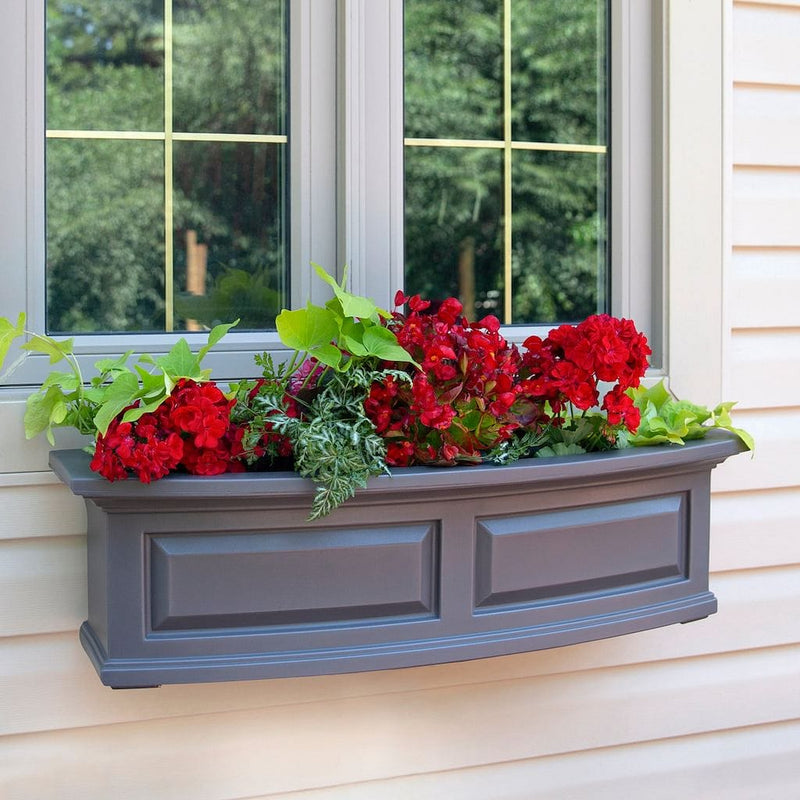 The Mayne Nantucket 3ft Window Box Planter, with a graphite finishmounted under a window and filled with colorful flowers.