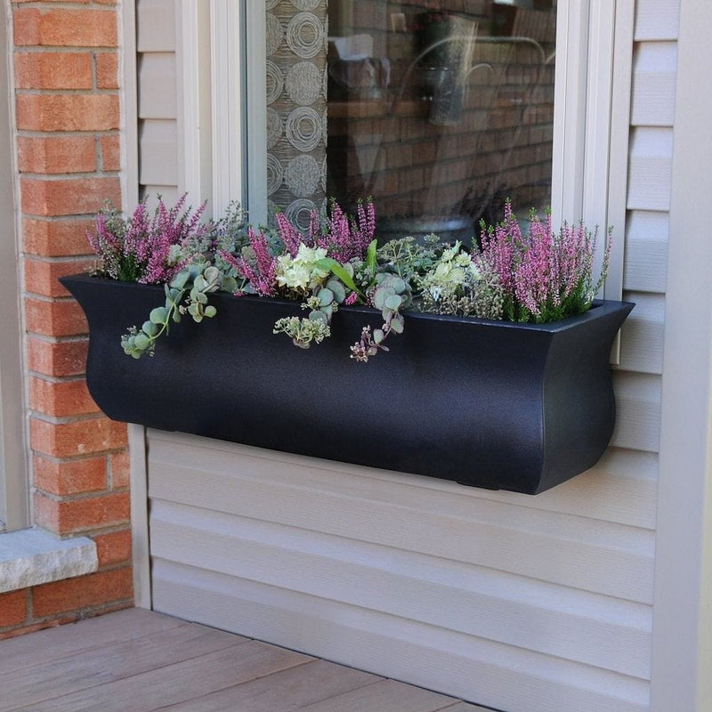 The Mayne Valencia 3ft Window Box Planter, with a black finish, mounted under a window and filled with colorful flowers.
