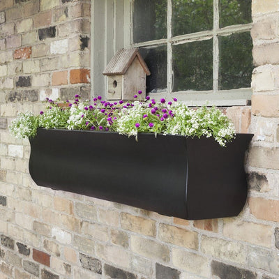 The Mayne Valencia 4ft Window Box Planter, in the black finish, planted with colorful flowers and hung under a brick framed window.