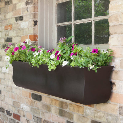 The Mayne Valencia 4ft Window Box Planter, in the espresso finish, planted with colorful flowers and hung under a brick framed window.