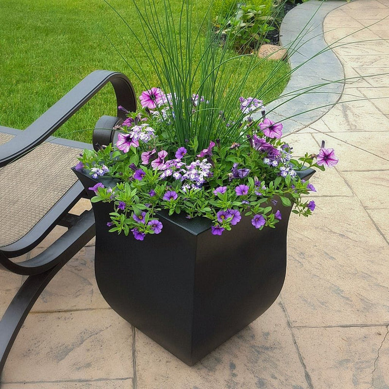 The Mayne Valencia Square Planter, in the black finish, planted with purple flowers and set out on a patio.