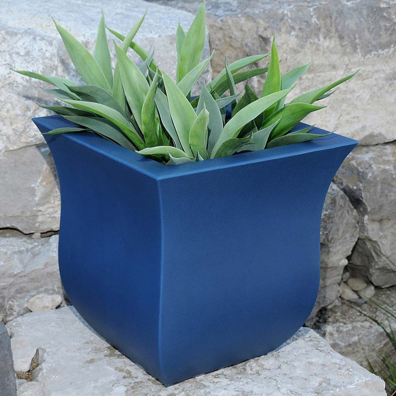 The Mayne Valencia Square Planter, in the neptune blue finish, filled with foliage and placed on a boulder in the garden,