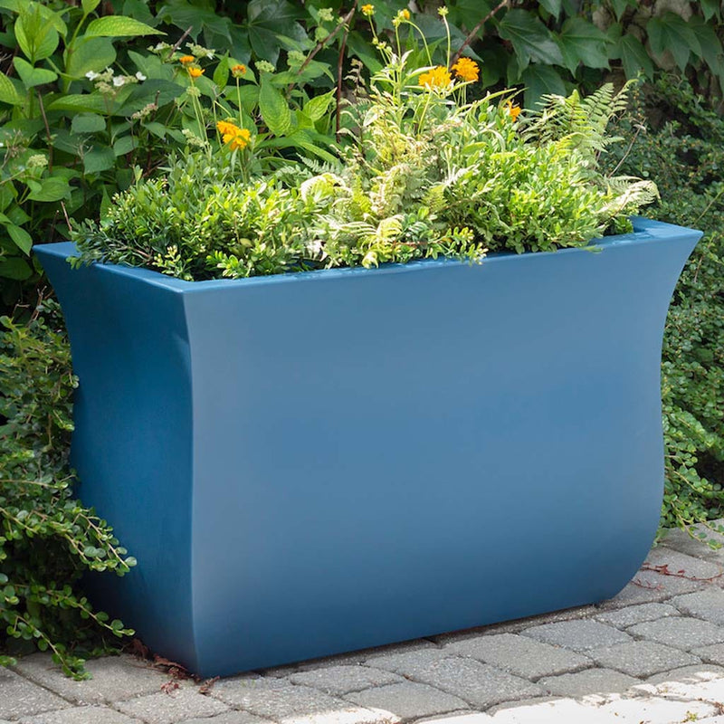 The Mayne Valencia Long Planter, in the neptune blue finish, planted with soft foliage and placed in the garden.