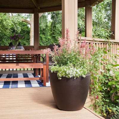 The Mayne Modesto Round Planter, in the espresso finish, placed on a deck in the sun filled with color annuals.