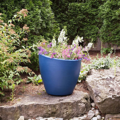 The Mayne Modesto Round Planter, in the neptune blue finish, filled with annual flowers and placed on a boulder in the garden.