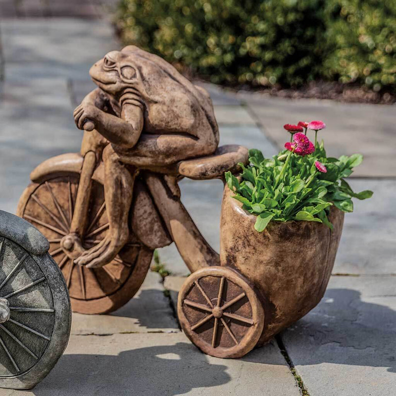 Campania International Tricycle Frog Planter Statue, set in the garden to add charm and character. The statue is shown in the Greystone Patina.