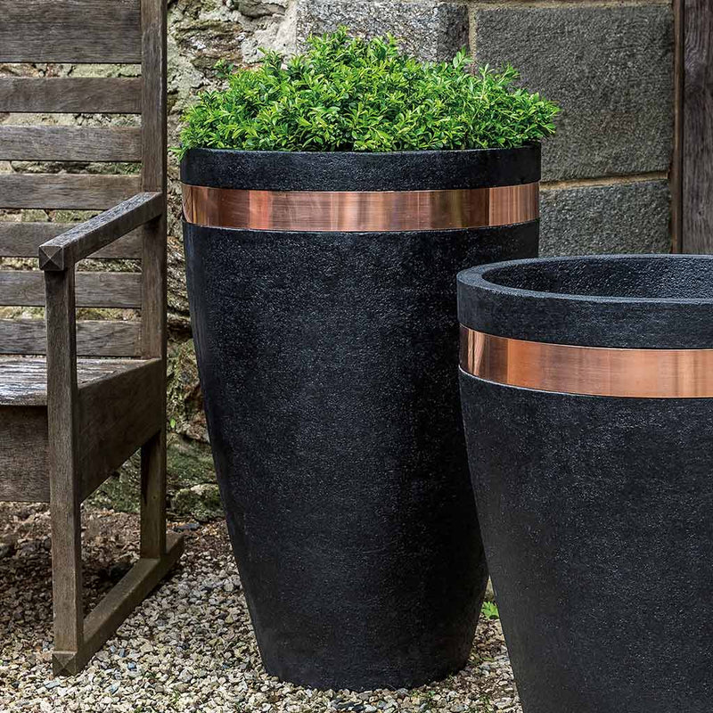 Campania International Moderne Tapered Tall Planter a dramatic combination of copper and stone, filled with plants and shown in the Nero Nuovo Patina. Made from cast stone.