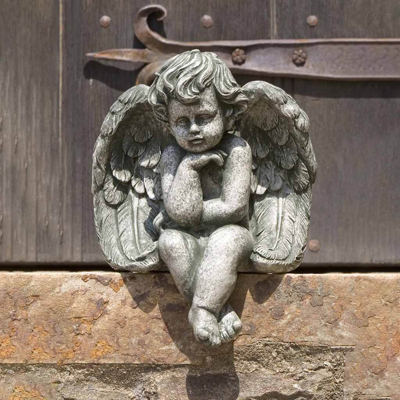 Campania International Small Sitting Cherub Statue placed in the garden. Religious garden statues, made of cast stone in a range of color options.