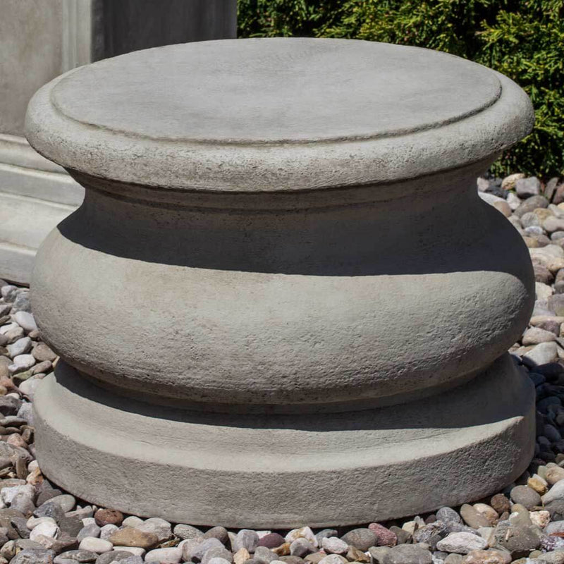 Campania International Low Round Plain Pedestal, set in the garden elevate a statue or planter. The pedestal is shown in the Greystone Patina.